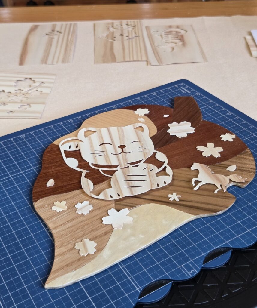 木象嵌の技法で制作した「木の葉っぱコースター」の上に飾られた、招き猫のシルエットの木の切り絵作品「春風を招いて」。