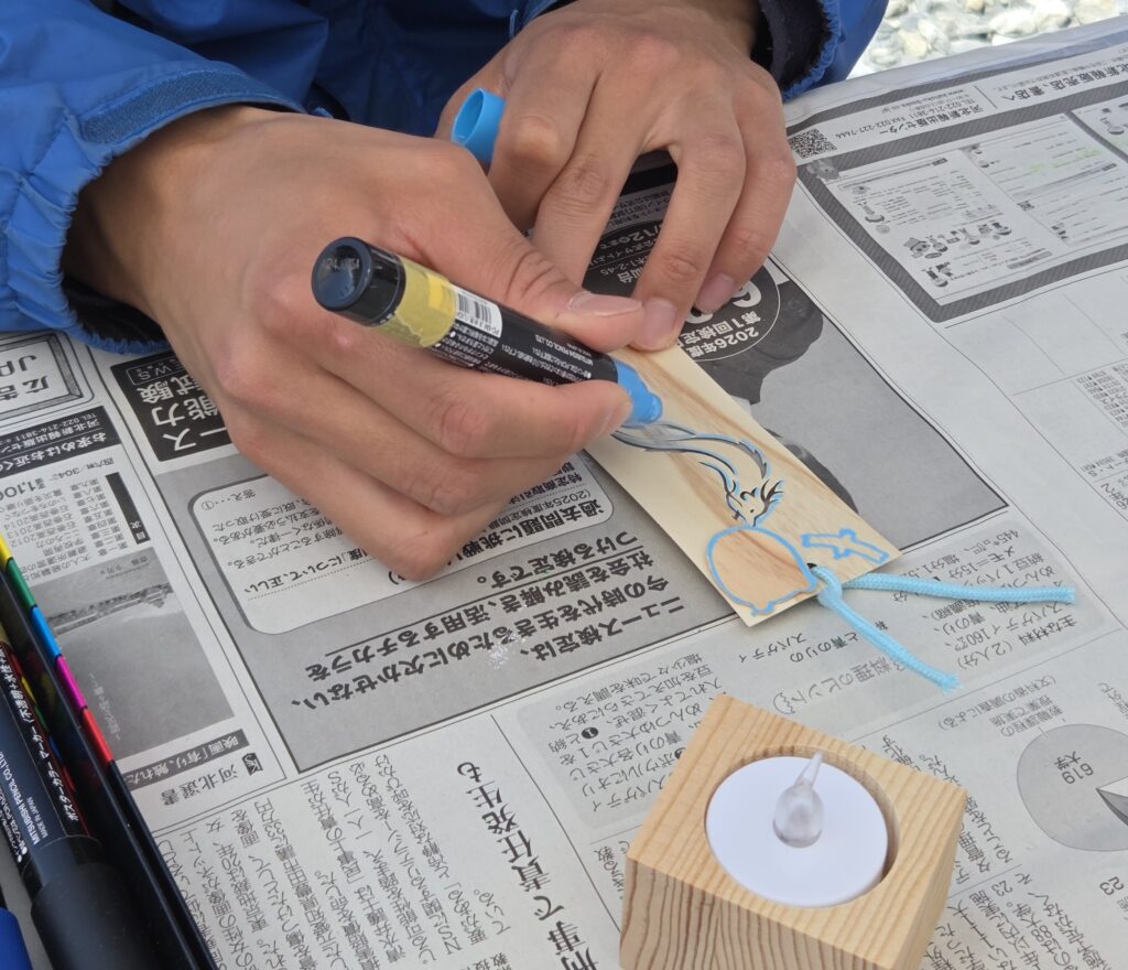 木の切り絵札お守りに、水色のペンで色を塗る人の手元。ワークショップに集中して作業している様子。